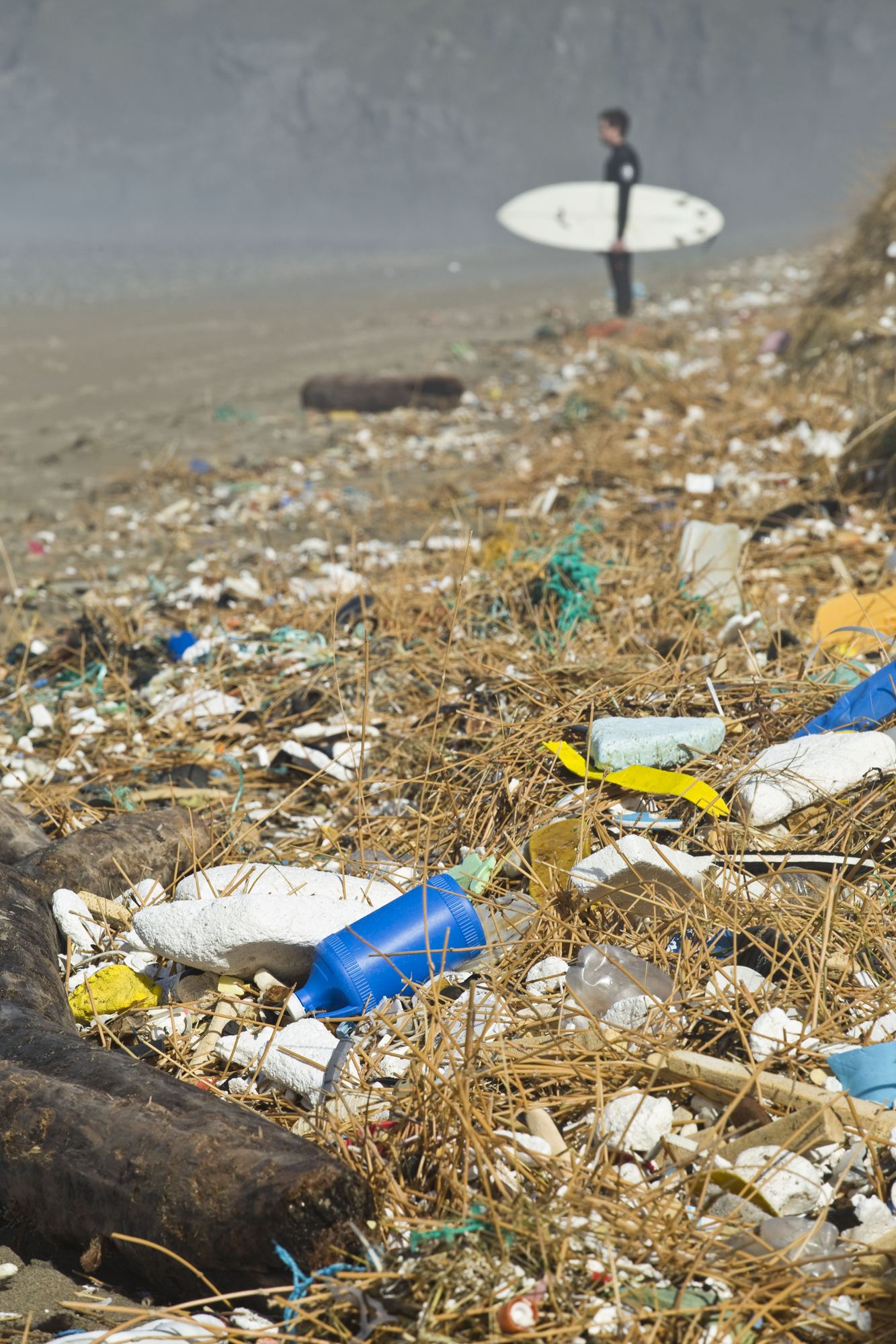 A surfer carrying a white board walks past a strandline thick with plastic bottles and debris