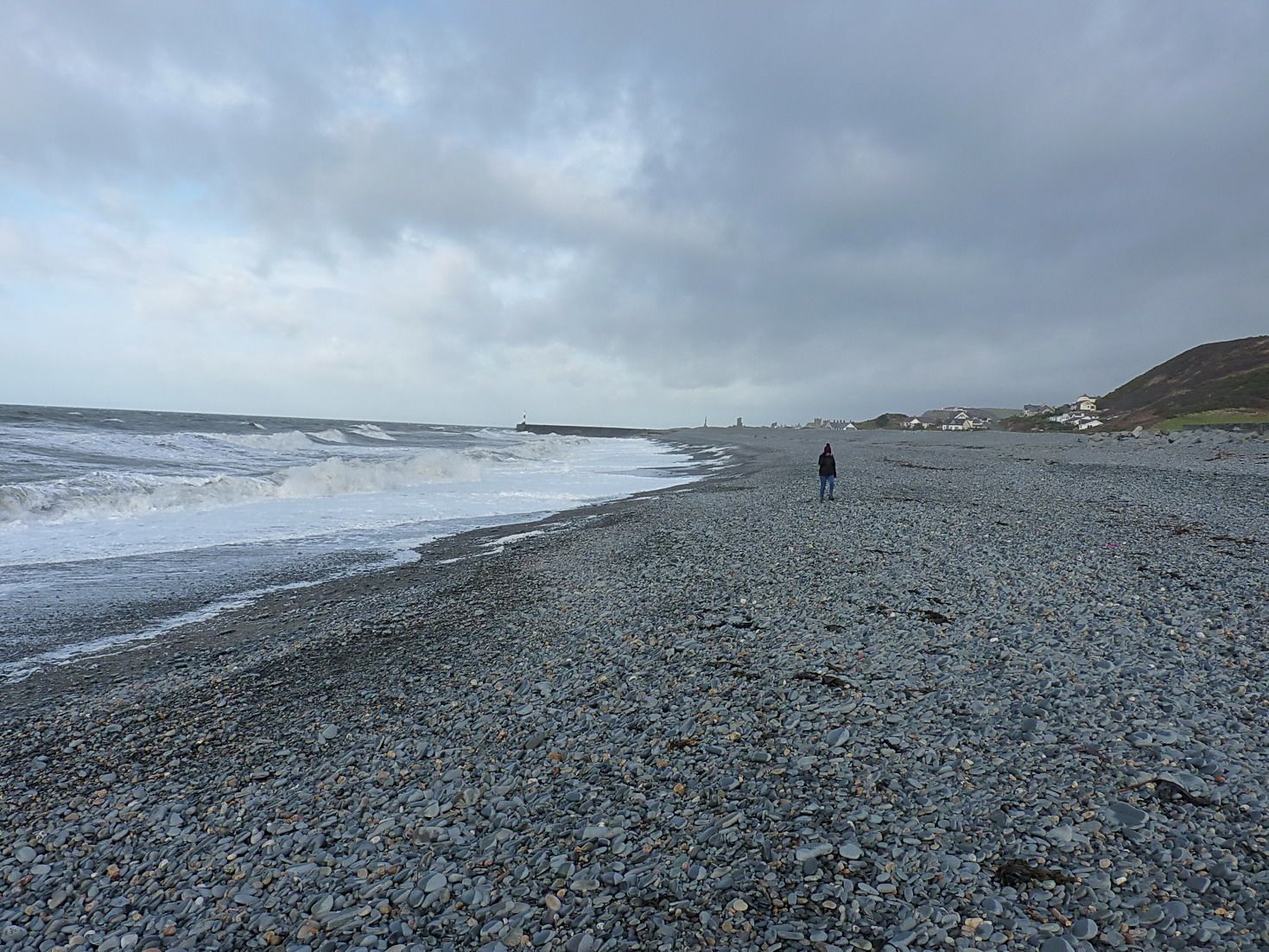 A lone walker far down a wide shingle beach with waves breaking on the left
