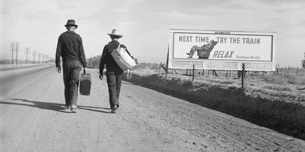 Two men walk past a billboard reading Next time try the train, Dorothea Lange, 1937