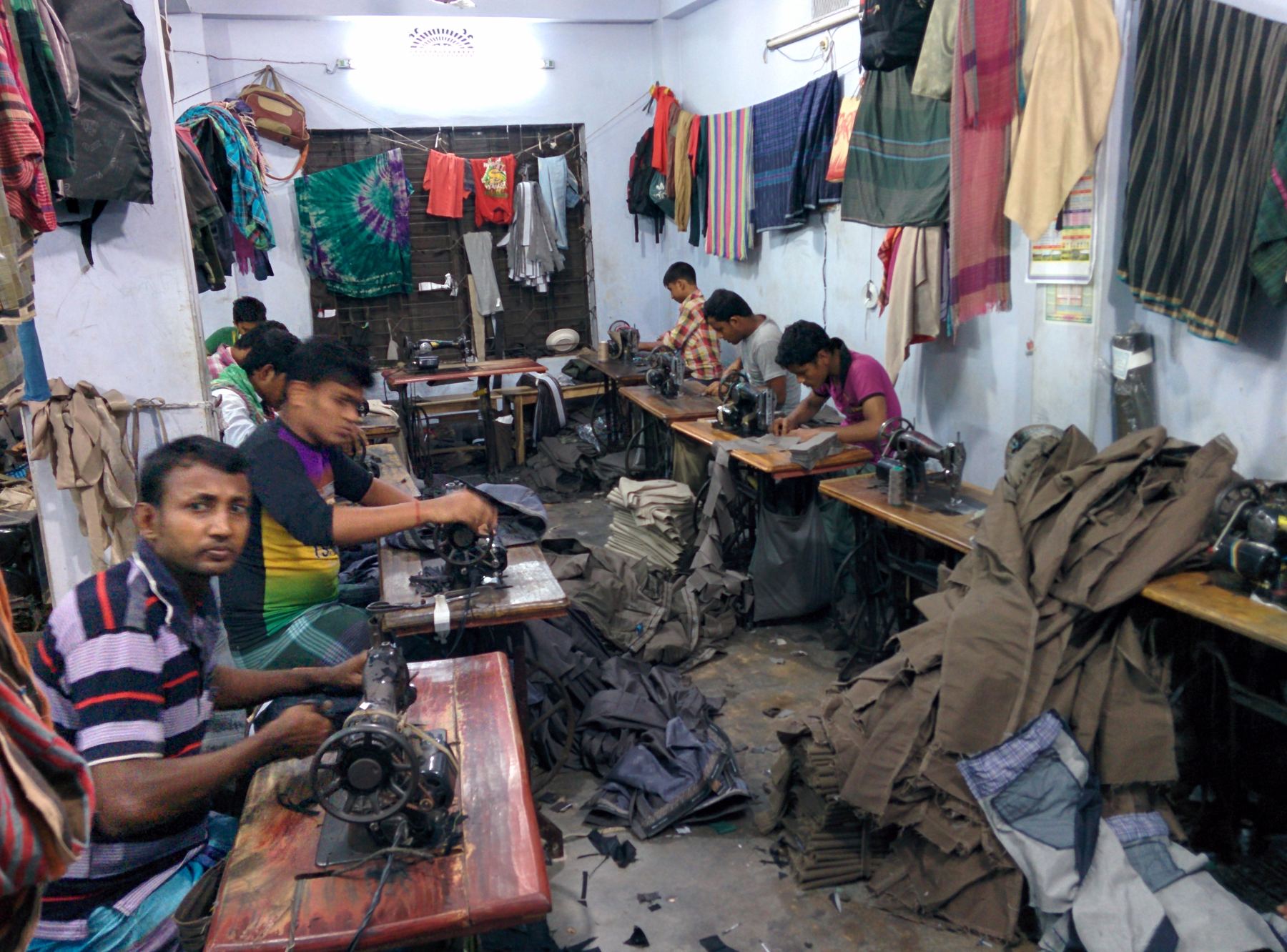 Men working at sewing machines in a crowded garment workshop in Dhaka, with clothes piled on tables and hanging from the ceiling