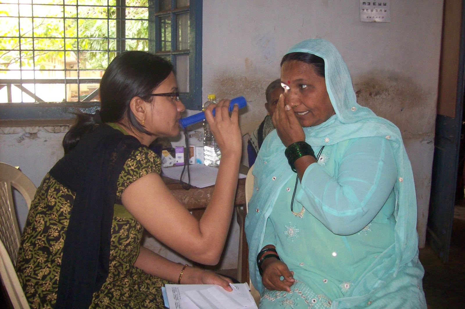 A woman in a turquoise headscarf covers one eye while a young woman holds a torch to check her vision