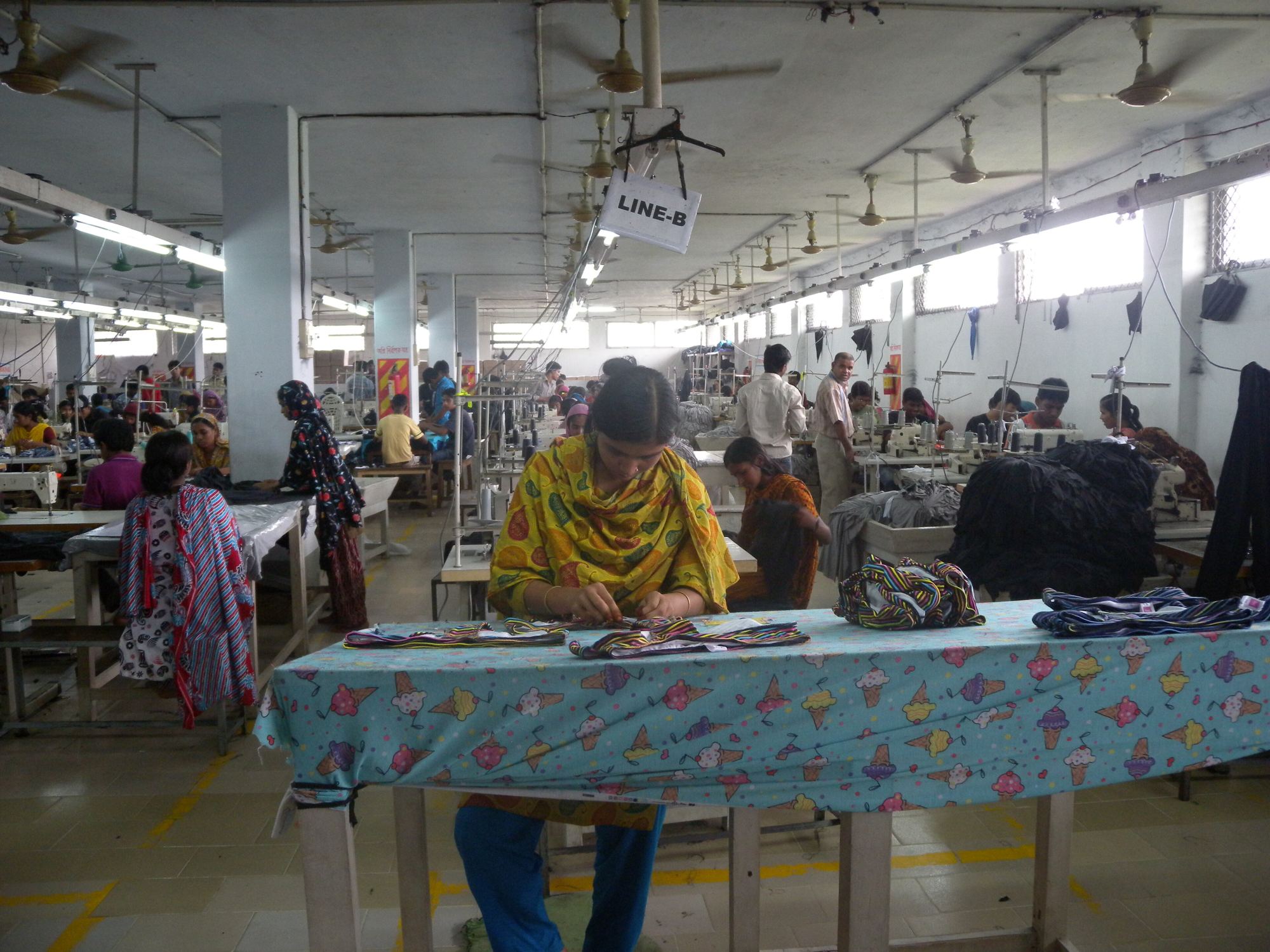 Rows of women working at sewing tables on a long garment factory floor in Bangladesh