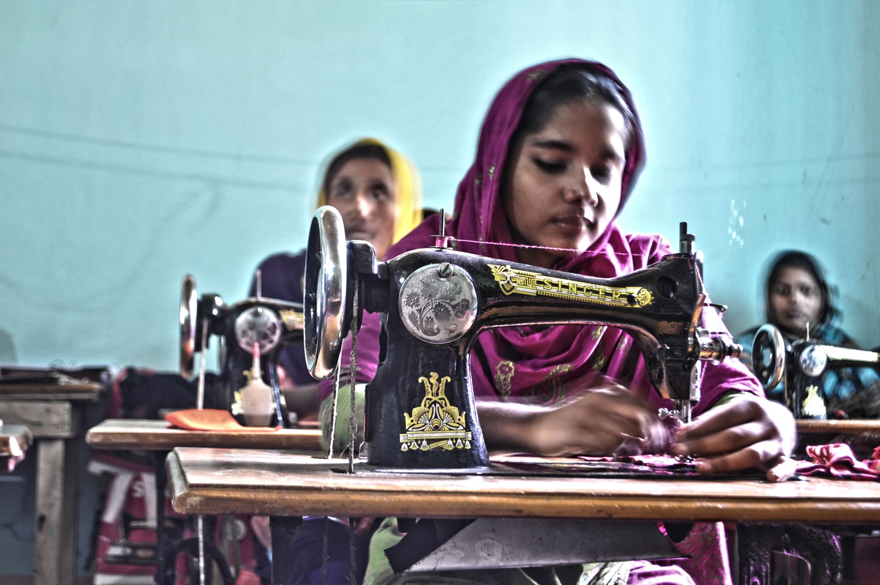 A young woman in a pink headscarf sewing at a black treadle sewing machine, with other women behind her