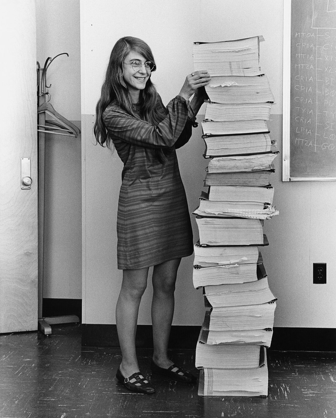 Margaret Hamilton standing next to a stack of printed Apollo program listings as tall as she is