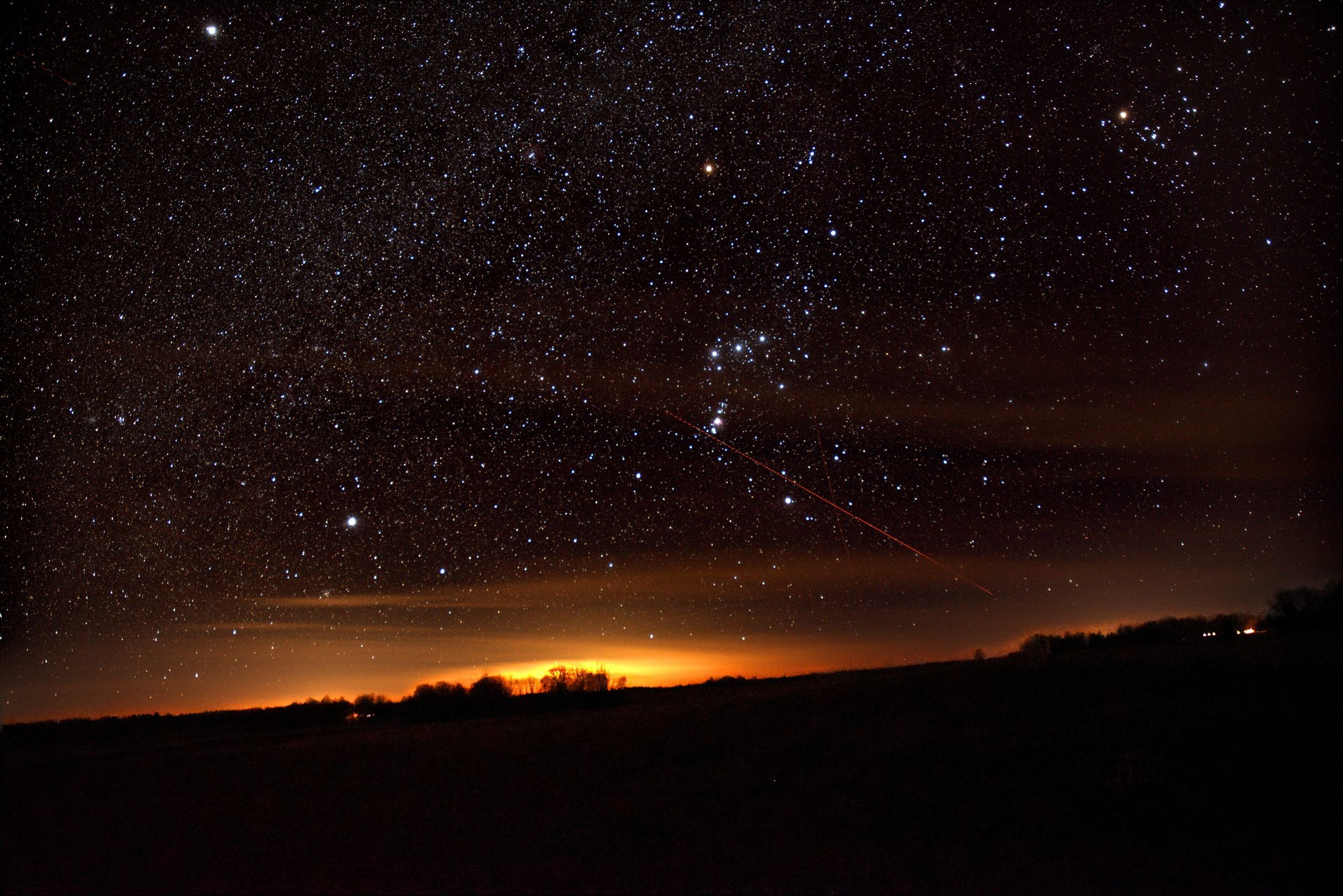 The constellation Orion in a starry sky, with an orange glow from town lights along the horizon