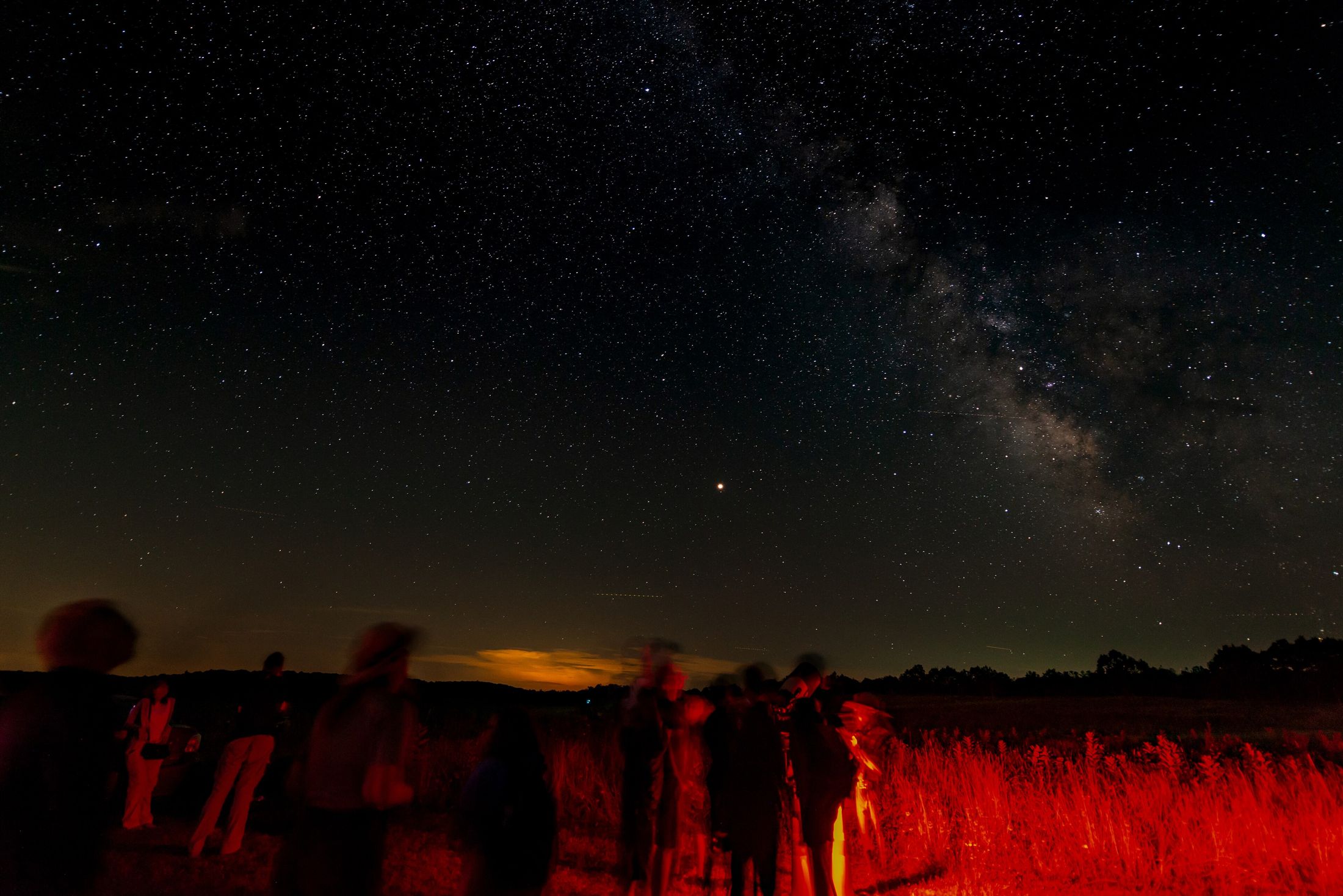 People with telescopes standing in a dark meadow under the Milky Way, lit by dim red torches