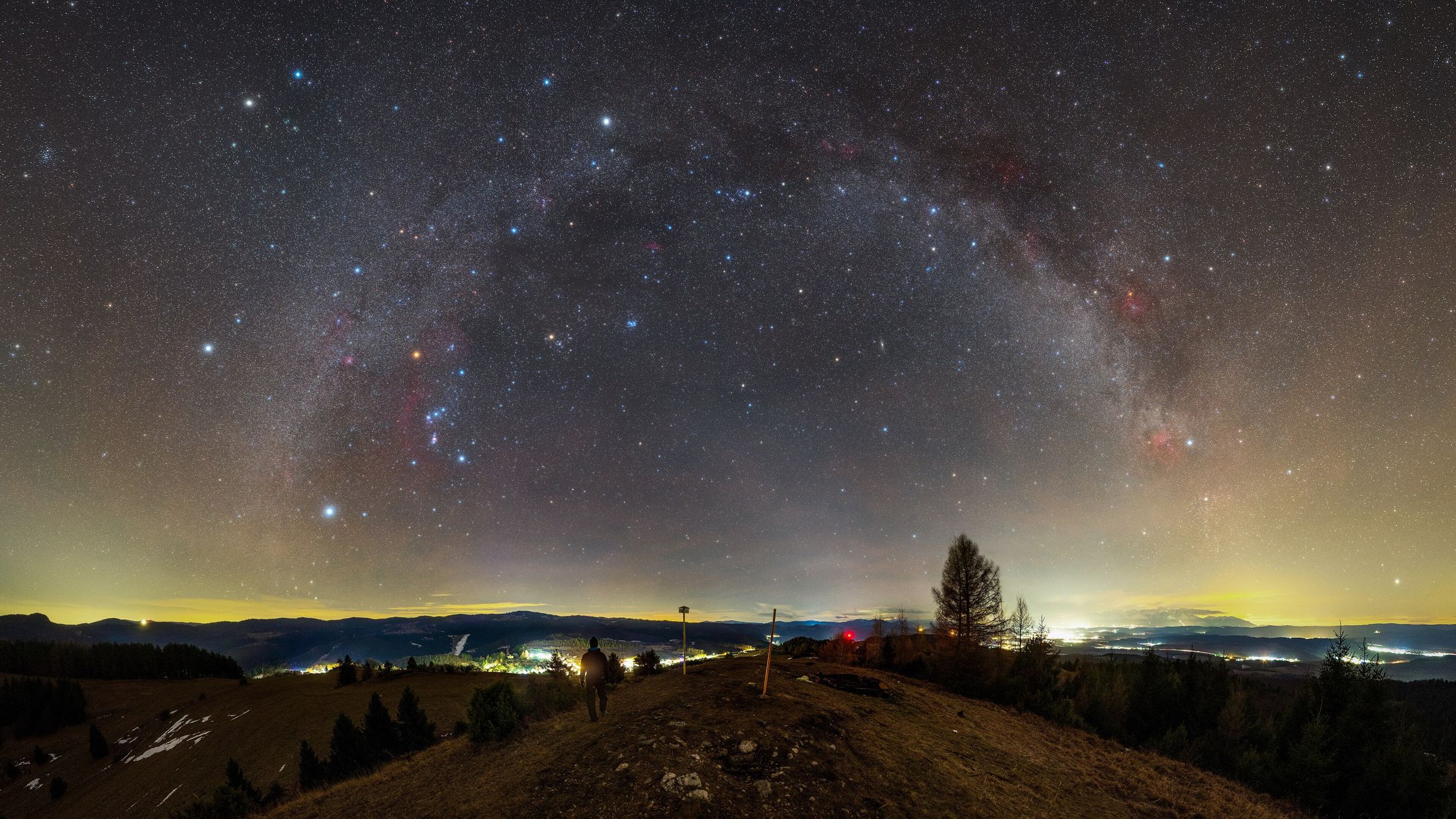The Milky Way arching over a hilltop in Slovakia, with the lights of towns glowing along the horizon