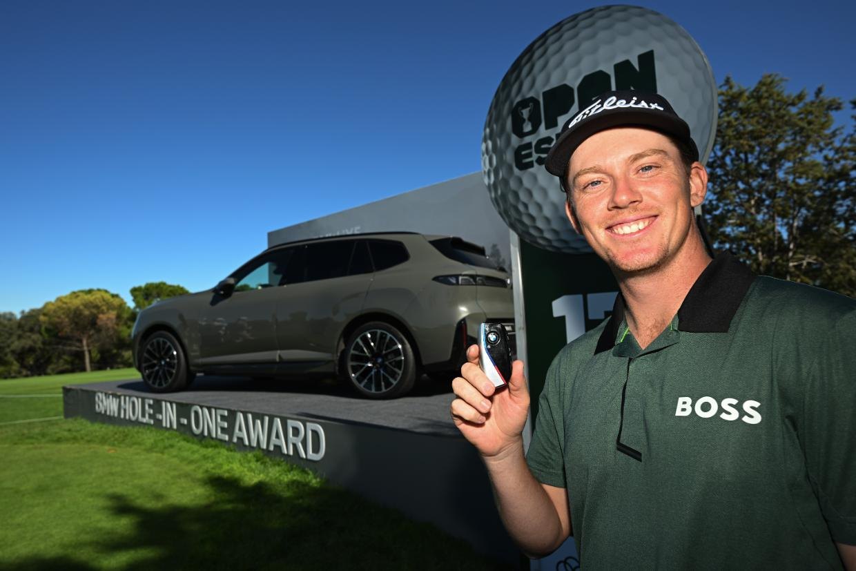 A smiling young golfer in a cap and green shirt holding up a car key, with an SUV on a stand marked BMW Hole-in-One Award behind him.