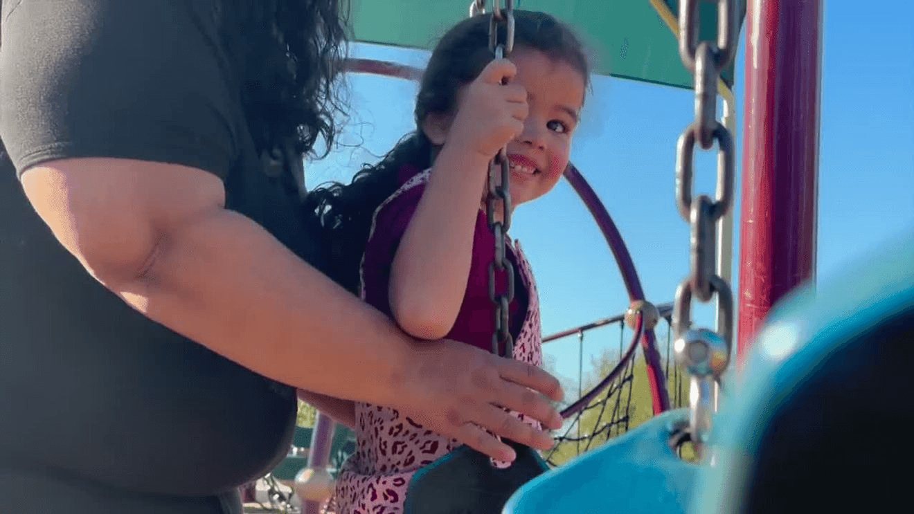 A small girl with dark hair in a pink leopard-print dress holding the chain of a swing and smiling, with a woman&rsquo;s arm beside her, under a blue sky.