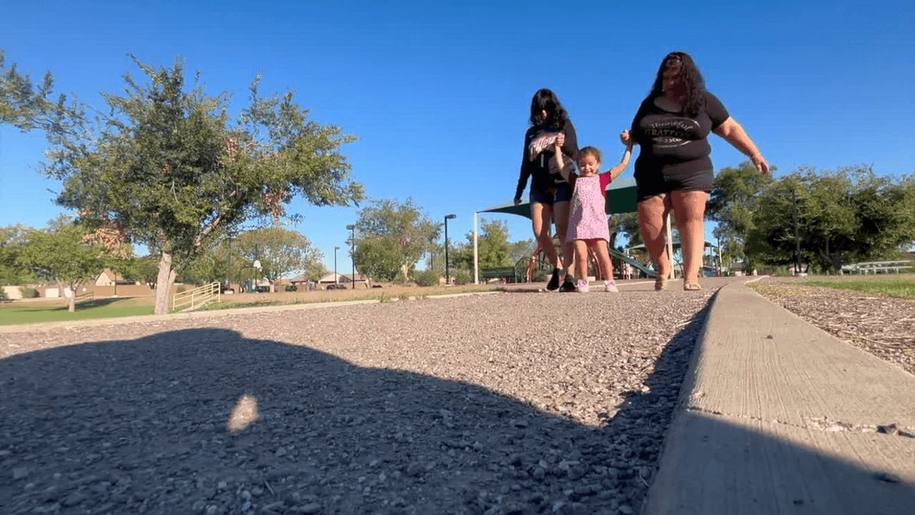 Alice walking along a park path holding hands with her grandmother and a teenage girl, trees and a playground shade behind them.