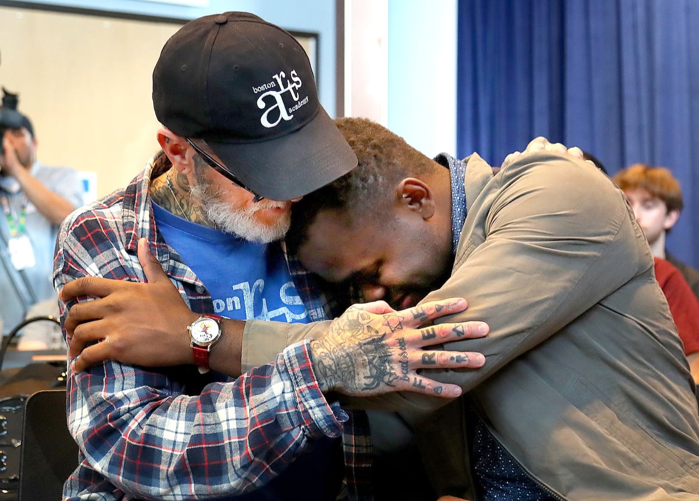 An older man with a white beard, tattoos and a Boston Arts Academy cap hugs a young man in an olive jacket, who rests his head on his shoulder.