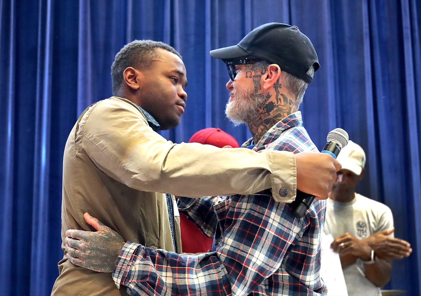On a stage with a blue curtain, the older man in the cap, holding a microphone, hugs the young man in the olive jacket.