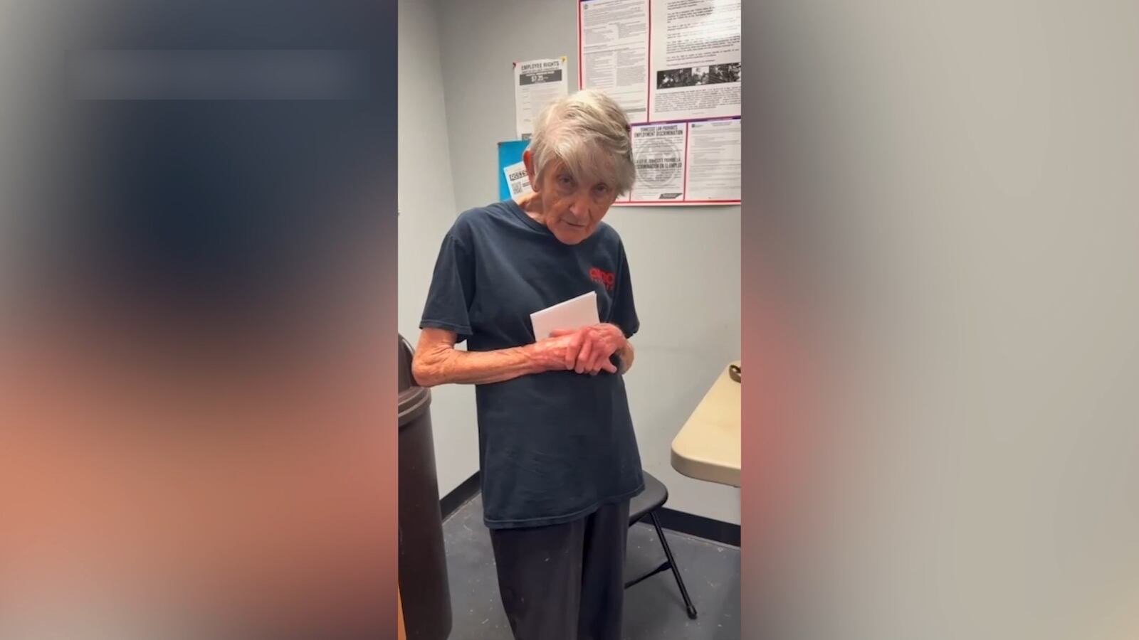 An older woman with short grey hair in a dark AMC T-shirt, holding a white envelope, in a staff room with notices on the wall.