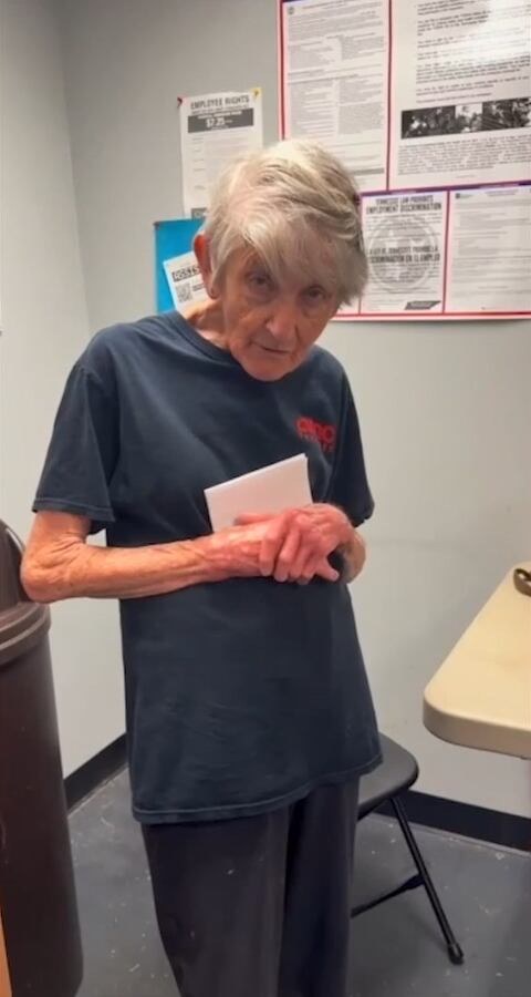 An older woman with short grey hair in a dark AMC T-shirt, holding a white envelope, in a staff room with notices on the wall.