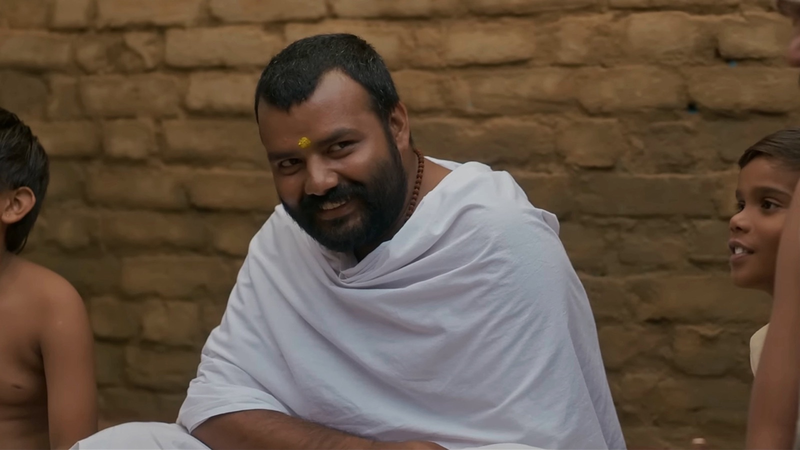 A bearded man in a white shawl laughs as he sits against a mud-brick wall between two young boys