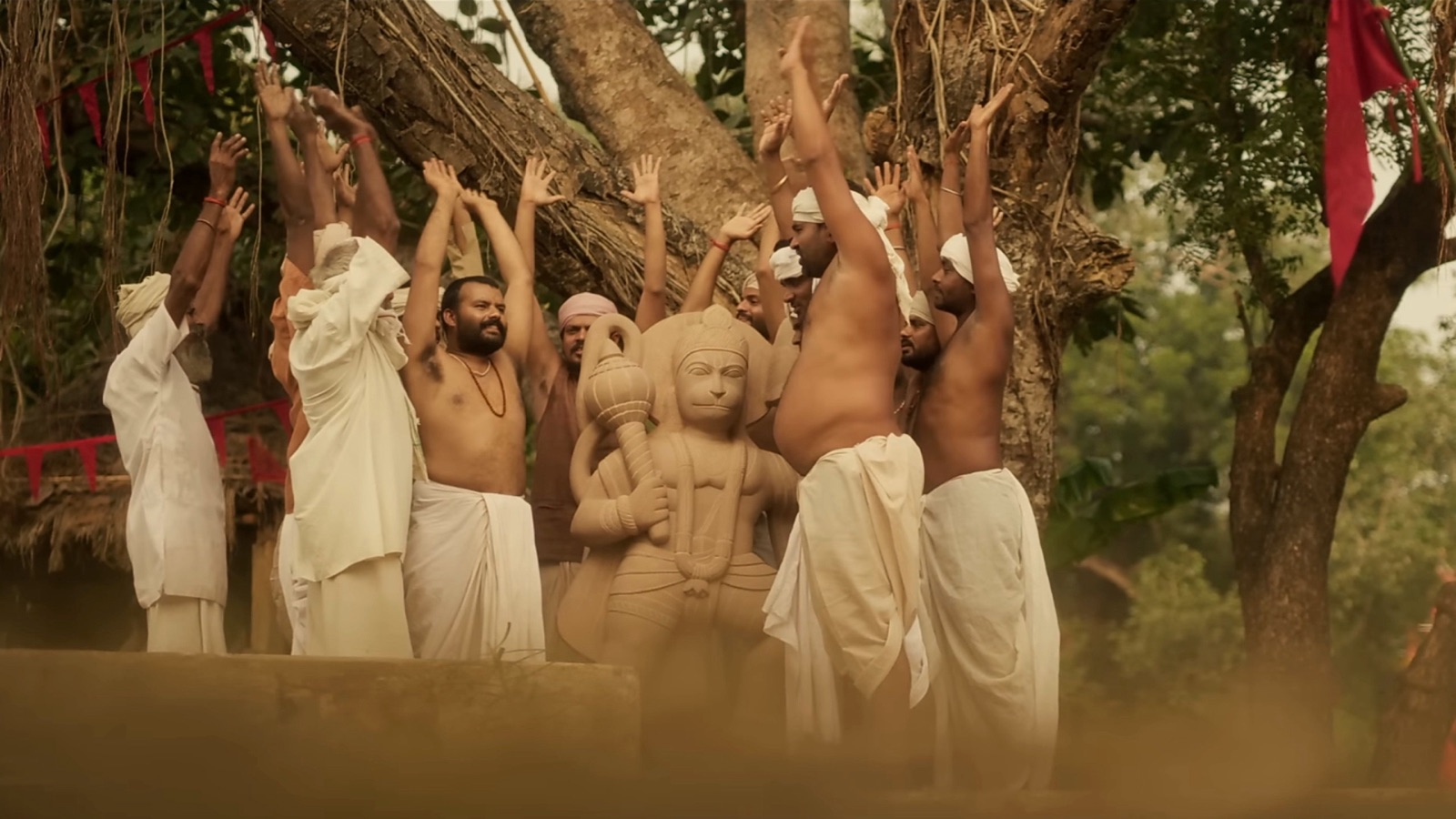 Men in white dhotis raise their arms around a stone statue of Hanuman under a large tree hung with red flags