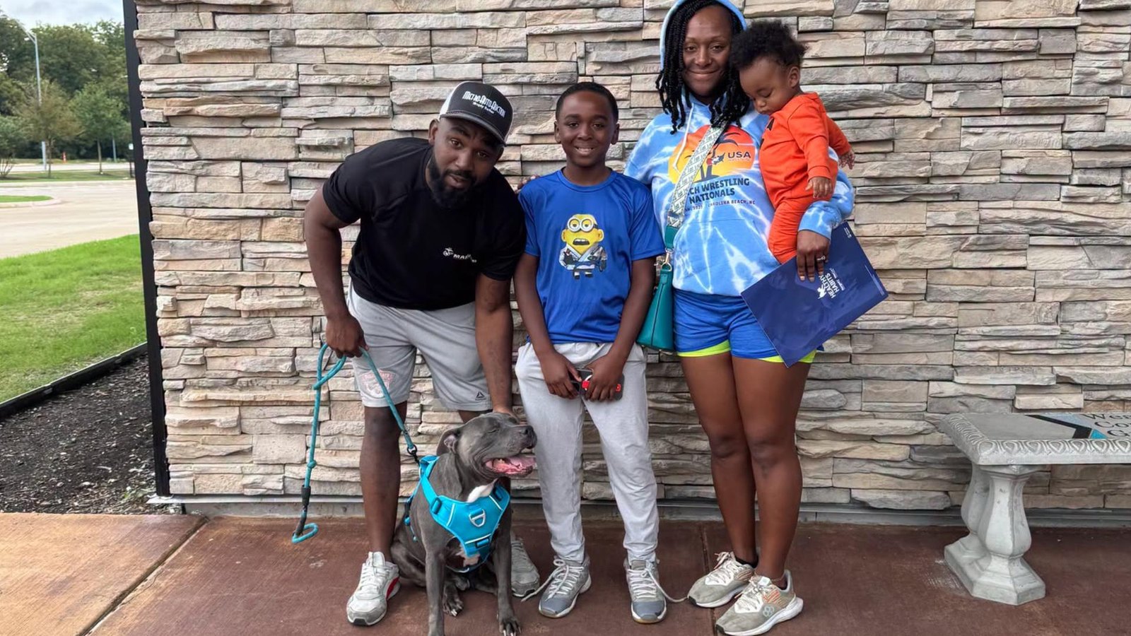 A grey dog in a blue harness sitting on a leash beside a family of four, a father, a boy, and a mother holding a toddler, in front of a stone wall.