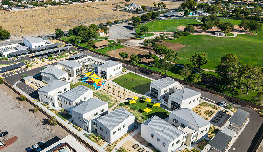 An aerial view of twelve white houses with grey pitched roofs arranged around a central green, with a playground under coloured shade sails, and a public park with a baseball diamond beyond