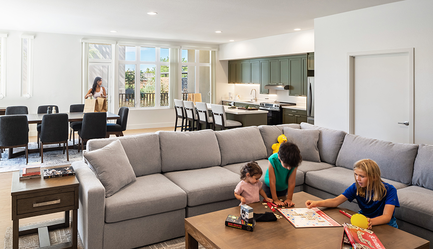 A living room in one of the homes: a large grey corner sofa, three children playing a board game on a low wooden table, an open kitchen with green cabinets behind, and an adult coming in through the door with a shopping bag
