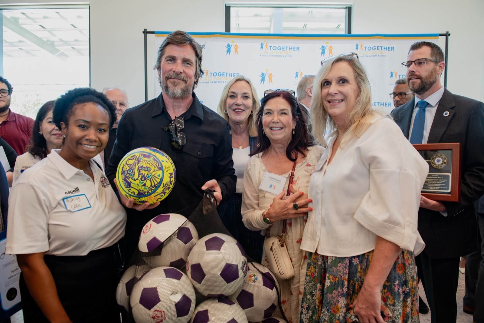 Christian Bale standing with several people in front of a Together California backdrop reading A Village for Brothers and Sisters, holding a signed football, with a bag of footballs in front of him and a framed plaque held at the right of the group