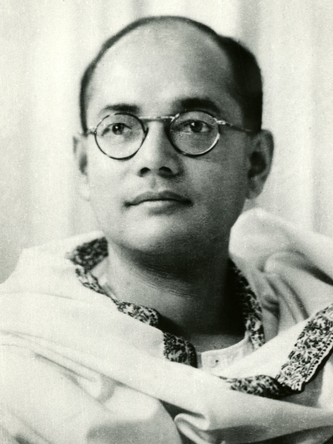A young Subhas Chandra Bose in round glasses and a white kurta with an embroidered neckline, looking slightly upward and away from the camera