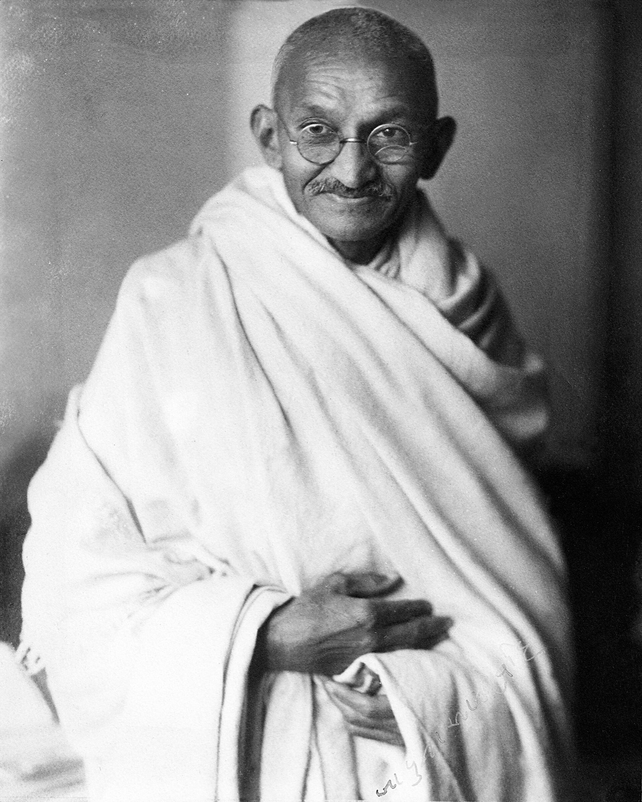Gandhi seated in a photographer's studio, wrapped in a white shawl, wearing round wire glasses, hands folded in his lap, looking at the camera with a slight smile