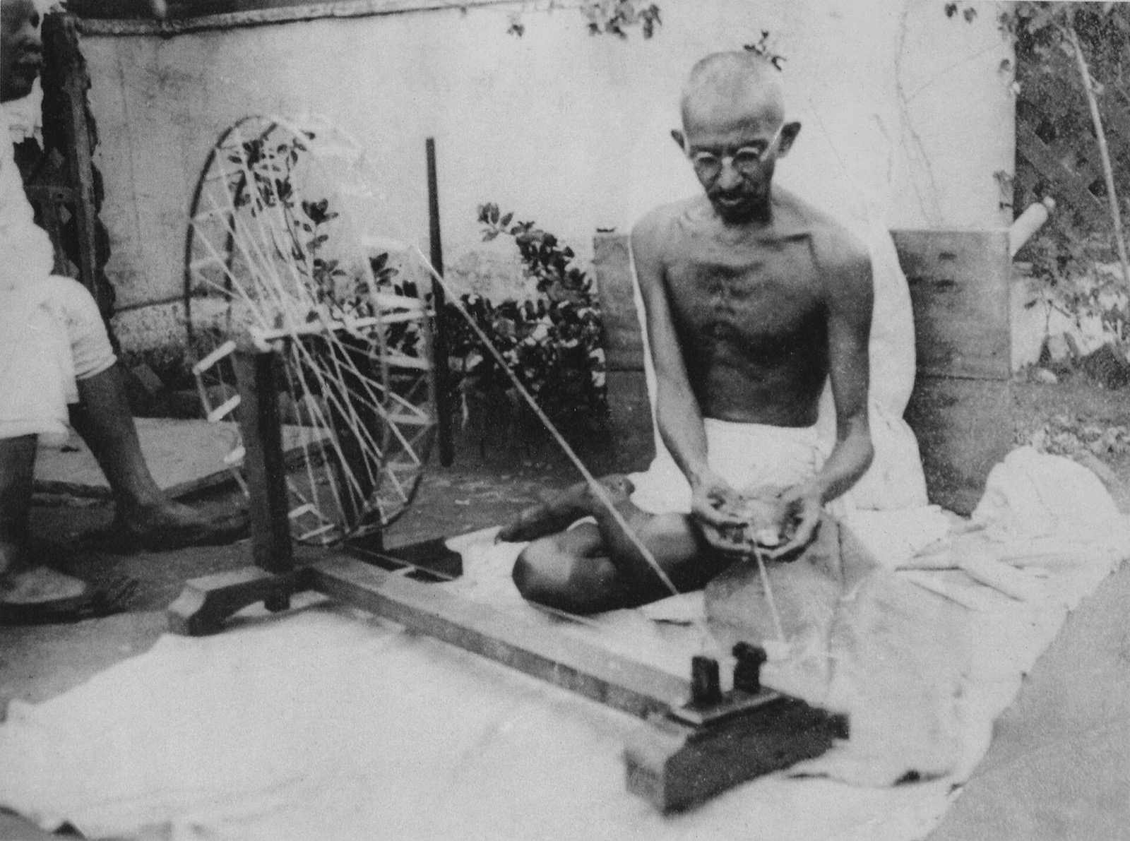 Gandhi sitting cross-legged on a white cloth on the ground outdoors, bare-chested in a dhoti, working at a wooden spinning wheel with another man standing behind him