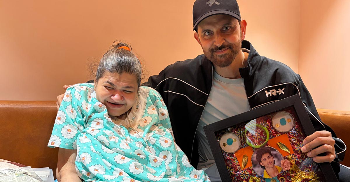 Hrithik Roshan in a black cap and jacket sitting on a sofa with his arm round Beeja, who wears a turquoise flowered nightdress and is laughing with her eyes closed, while he holds up a framed collage with his photograph at its centre