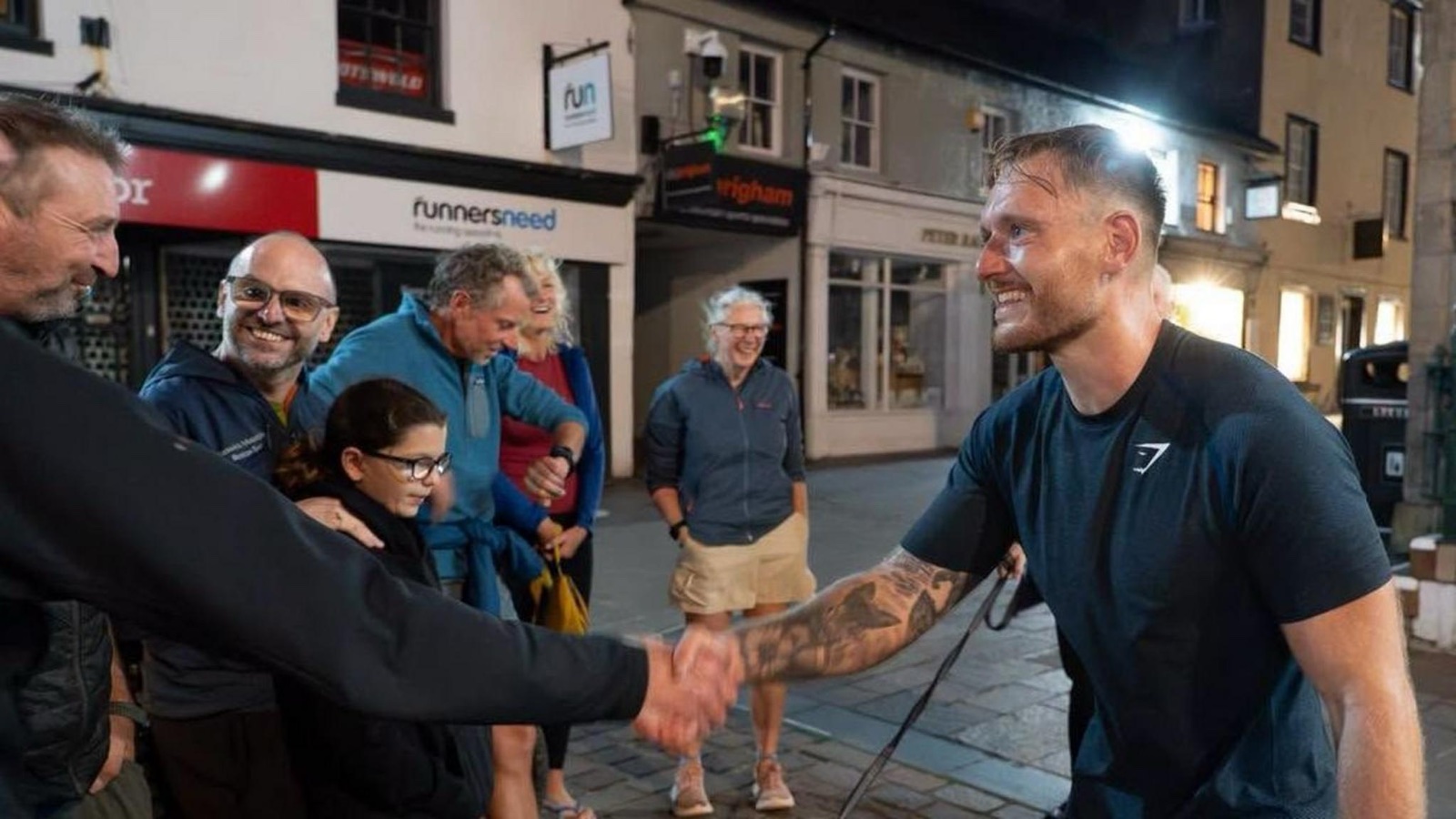 Tommy Price, sweating and grinning in a dark T-shirt, shaking hands with a man in the street in Keswick at night while a small crowd looks on and smiles