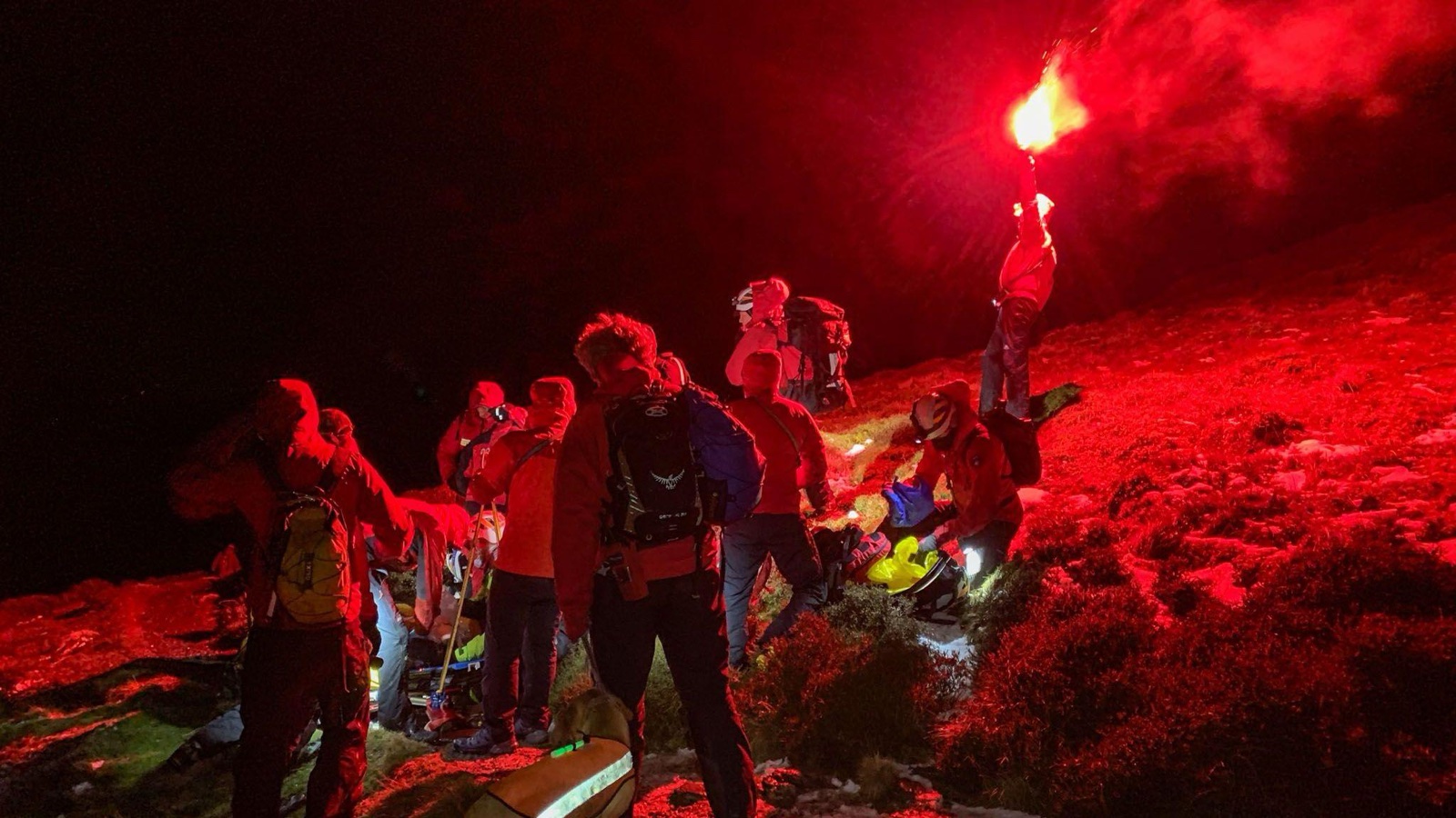 A mountain rescue team in red jackets gathered around a stretcher on a dark fellside, one of them holding a red flare above their heads