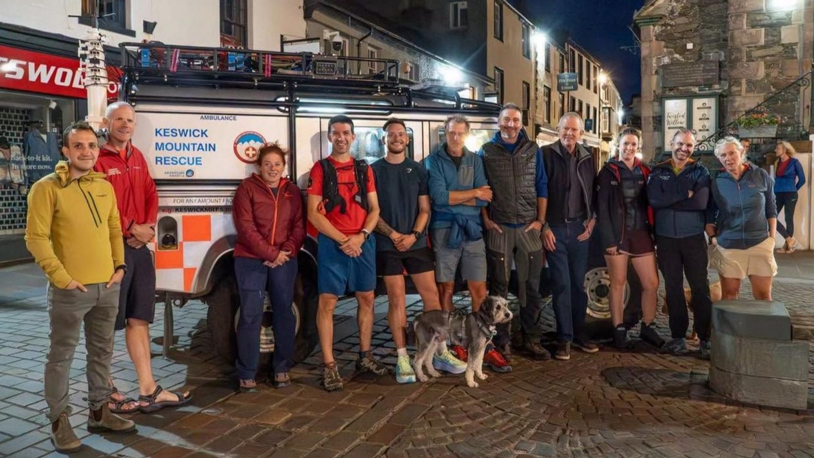 The Keswick Mountain Rescue team standing in a line in front of their ambulance in Keswick town centre at night, with a dog sitting at their feet