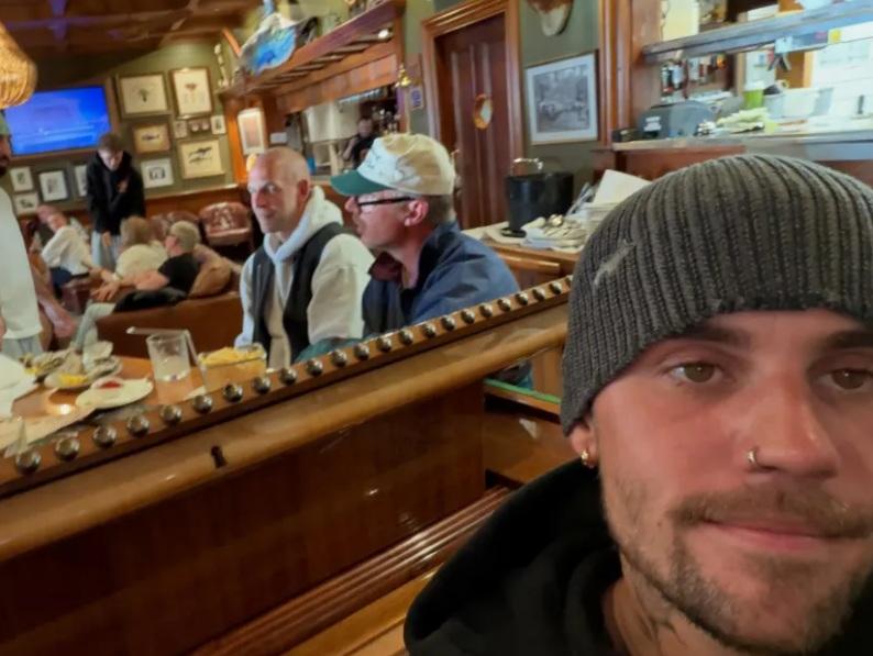 Justin Bieber in a grey beanie takes a selfie at a piano, with the restaurant's wooden bar and diners behind him.