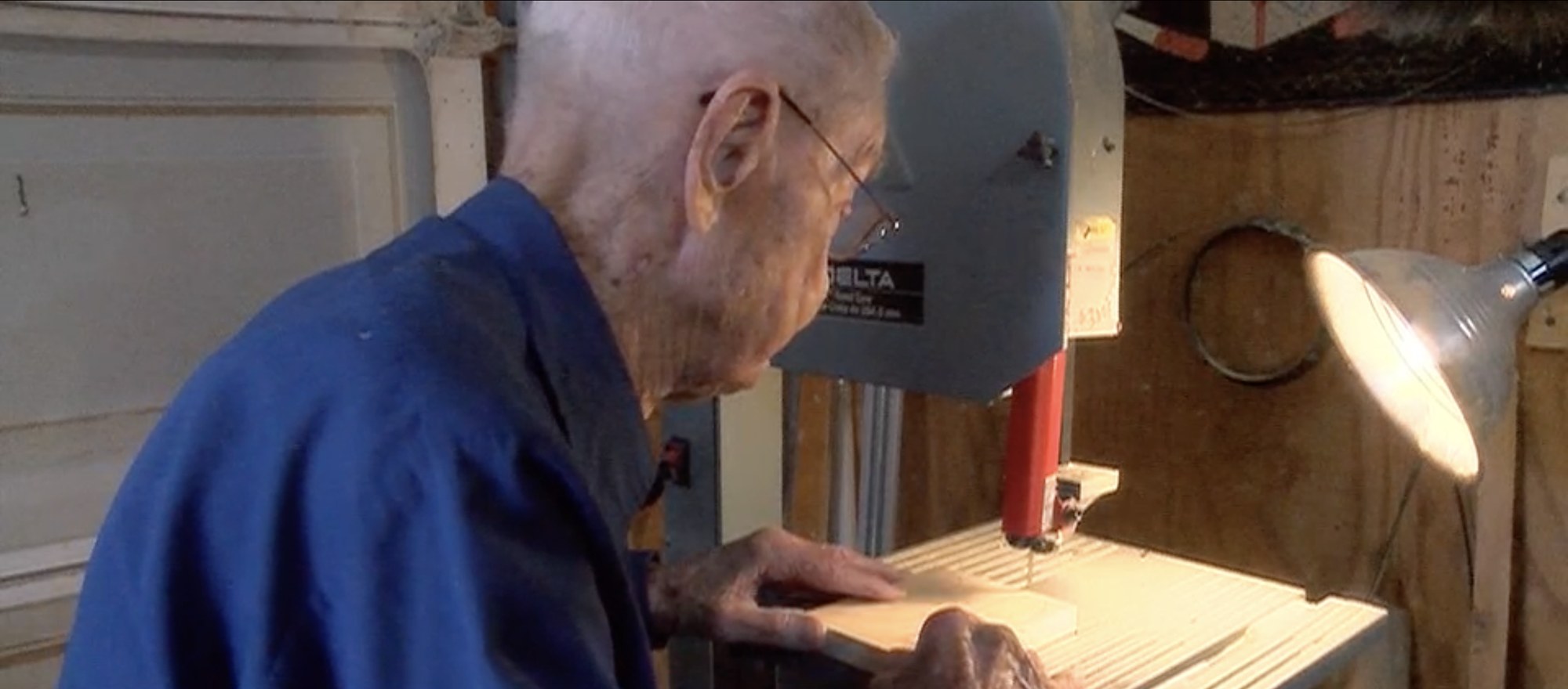 Bill Covington, aged one hundred, bent over a blue Delta bandsaw in his workshop, wearing a blue shirt and wire glasses, both hands flat on a pale board as he feeds it towards the blade, an angled lamp lighting the table