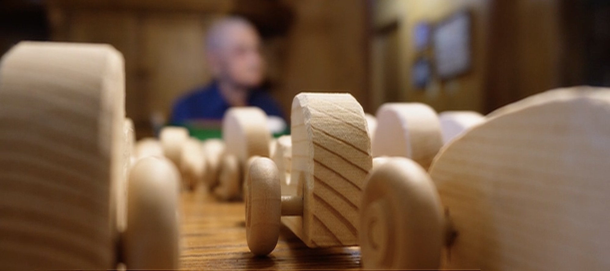 A row of finished pale wooden toy cars standing on a dark polished table, their round wooden wheels in focus in the foreground, with Bill Covington out of focus behind them in his blue shirt