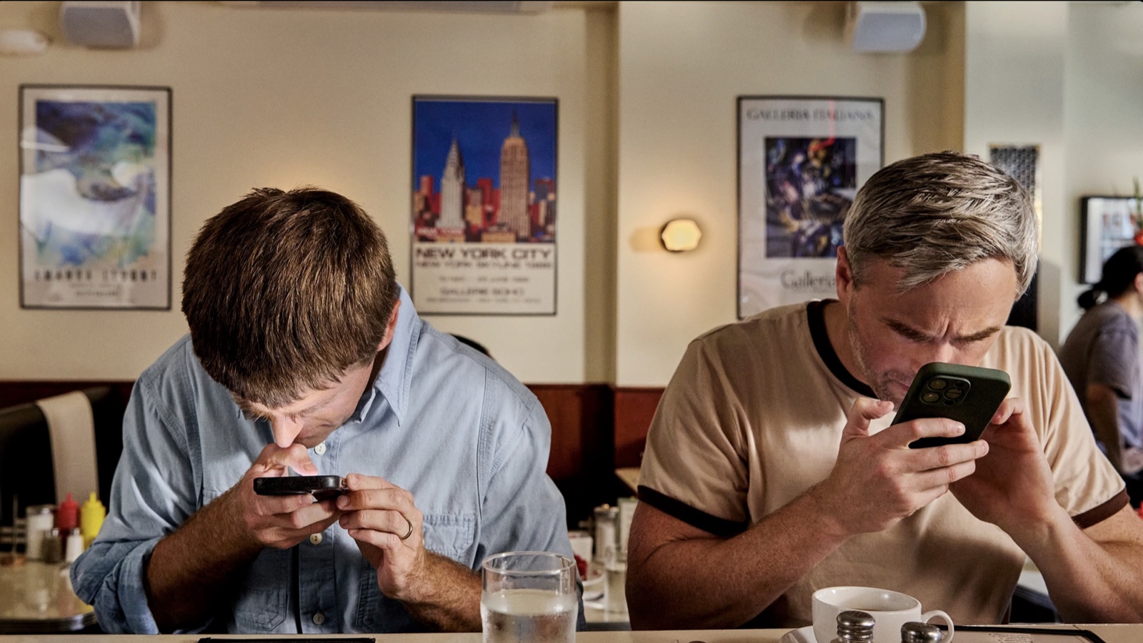 Two men sit side by side in a diner booth under framed New York posters, each bent low over a phone held close to his eyes