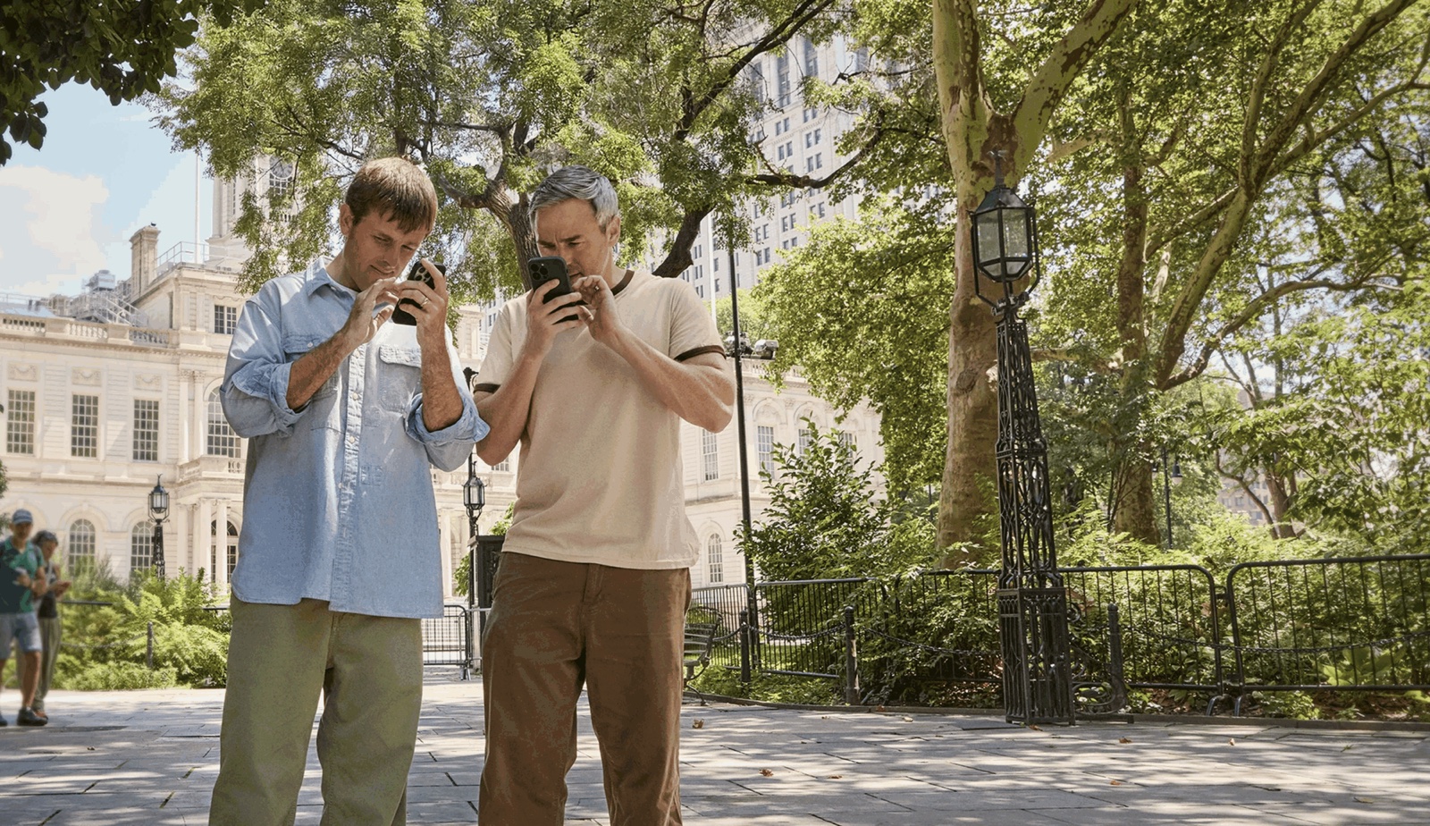 Two men stand on a path outside a white civic building in New York, each holding a phone a few inches from his face and reading it closely