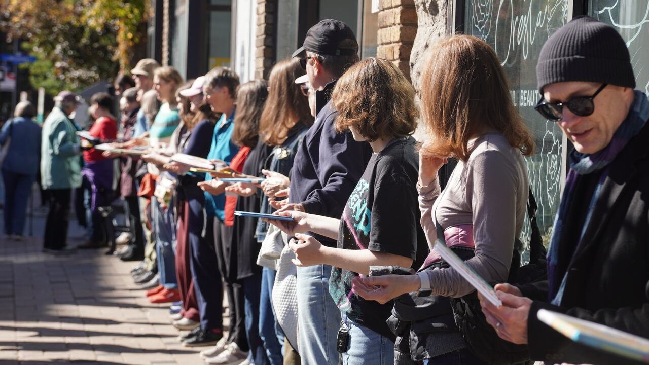 A long line of people standing along a city pavement in autumn sunshine, passing books from hand to hand.
