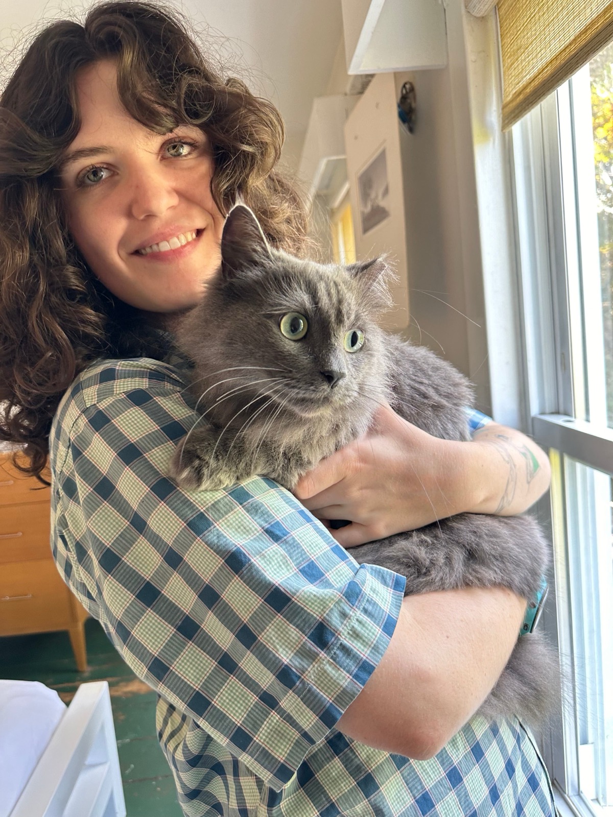 A smiling young woman with curly dark hair, in a checked shirt, holding a long-haired grey cat in her arms by a window.