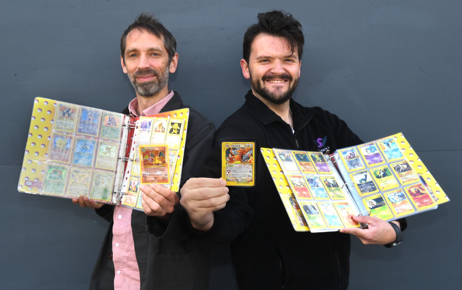 Two men against a grey wall holding open ring binders full of Pokémon cards in plastic sleeves, one of them also holding up a single Charizard card