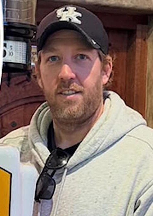 Alastair Cass, a man in his forties with a short beard, wearing a dark cap and a grey hooded top, sunglasses hanging at his collar, standing in a doorway