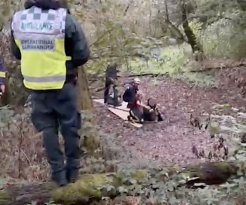 Crews working in a muddy wooded gully in Devon: an ambulance operational commander in a hi-vis jacket watching from higher ground while two rescuers kneel on boards laid across the mud beside the woman, whose face is obscured
