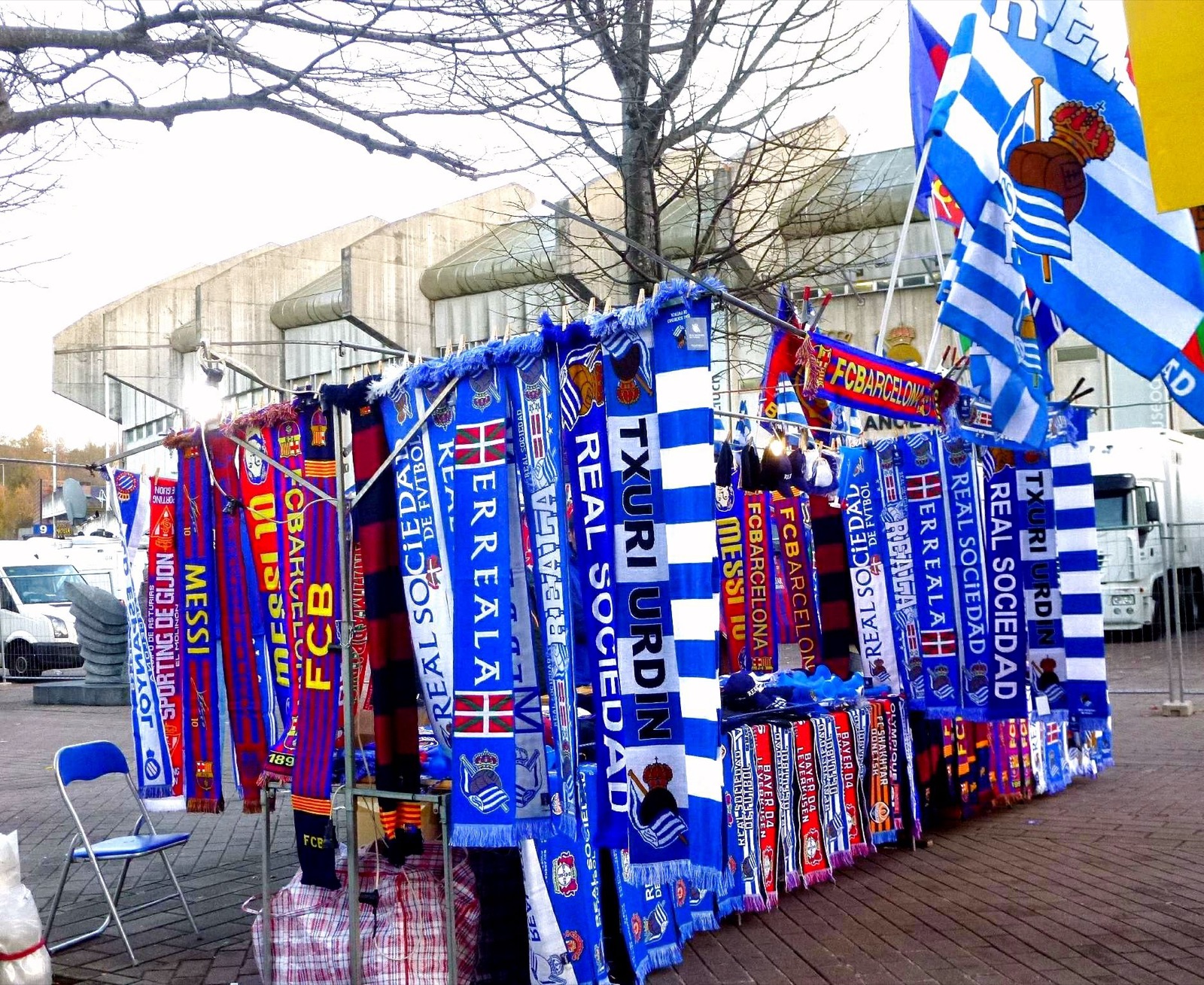 A stall of blue and white Real Sociedad scarves and flags hung on a rail outside the concrete stands of the Anoeta, with a folding chair beside it