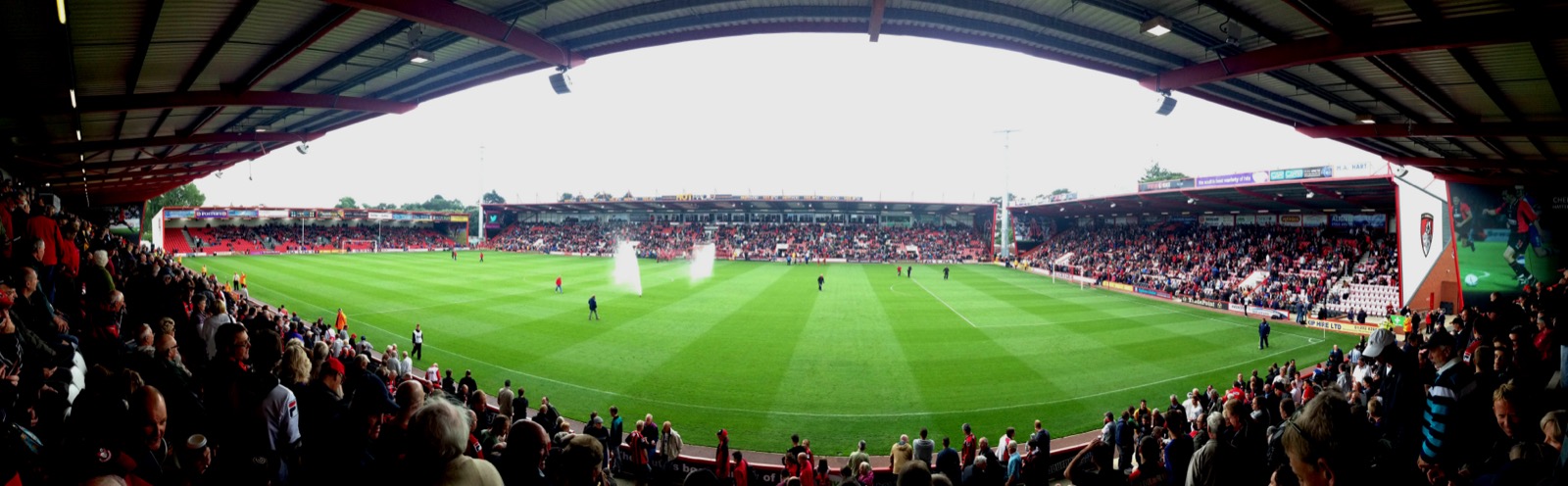 A wide view of Dean Court from the east stand, three low red-roofed stands around a green pitch with sprinklers running before a match, supporters in red and black in the foreground