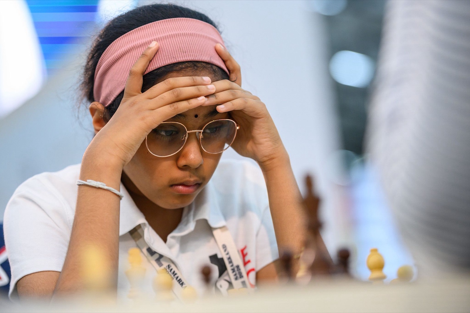 Bodhana Sivanandan holds her head in both hands as she studies the board during a game, wearing glasses, a pink headband and a white England shirt