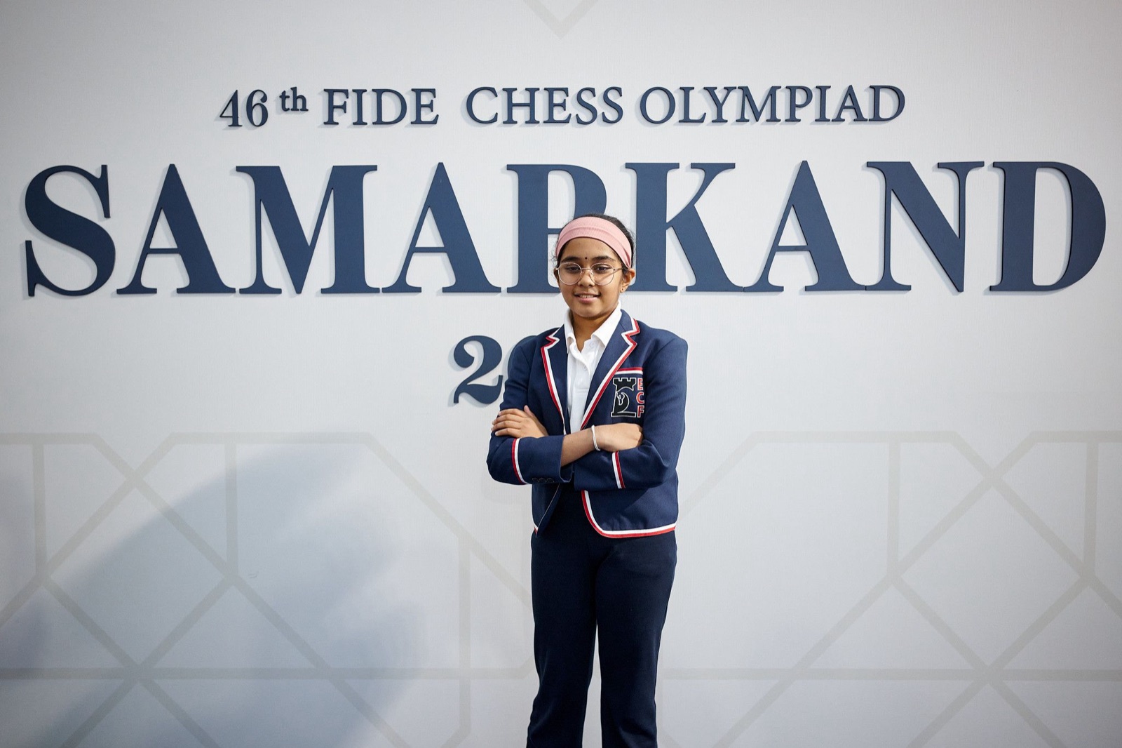 Bodhana Sivanandan, in round glasses, a pink headband and a navy England blazer, stands with her arms folded in front of a large sign reading 46th FIDE Chess Olympiad Samarkand
