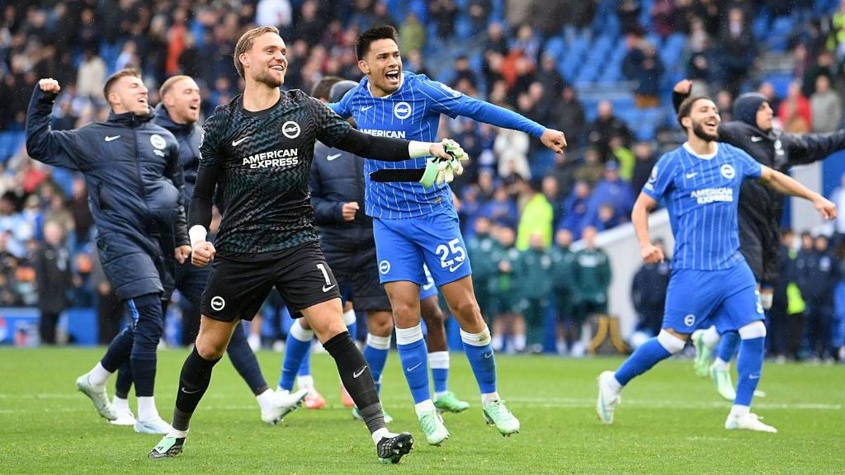 Brighton goalkeeper Bart Verbruggen, in a black kit with the number 1, celebrates on the pitch with teammates in blue and white after the win over Crystal Palace, with fans in the stand behind them.
