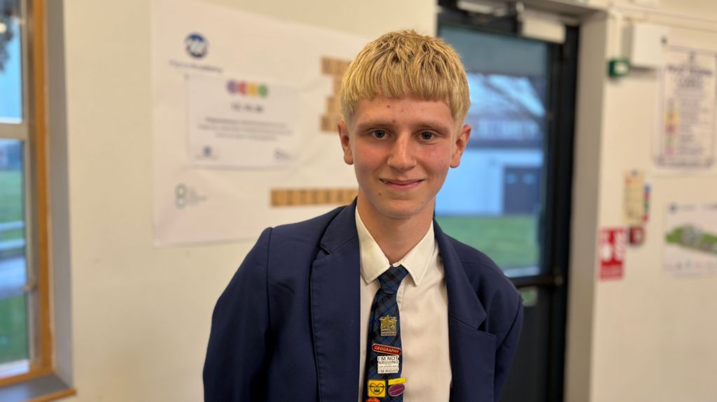 A teenage boy with blond hair in a navy school blazer and tie stands in a school corridor, smiling.