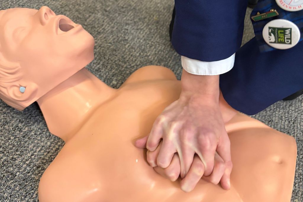A pair of hands in a navy blazer sleeve press on the chest of a CPR training dummy lying on a carpet.