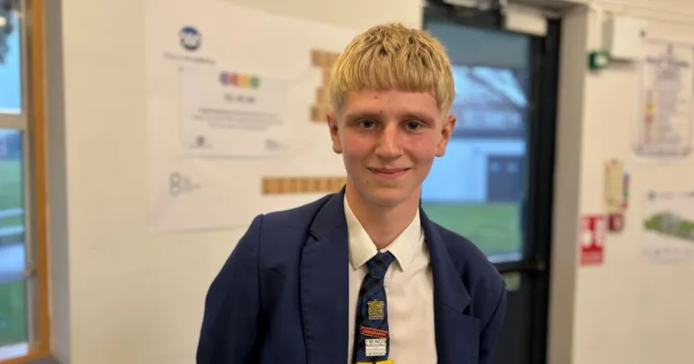 A teenage boy with blond hair in a navy school blazer and tie stands in a school corridor, smiling.