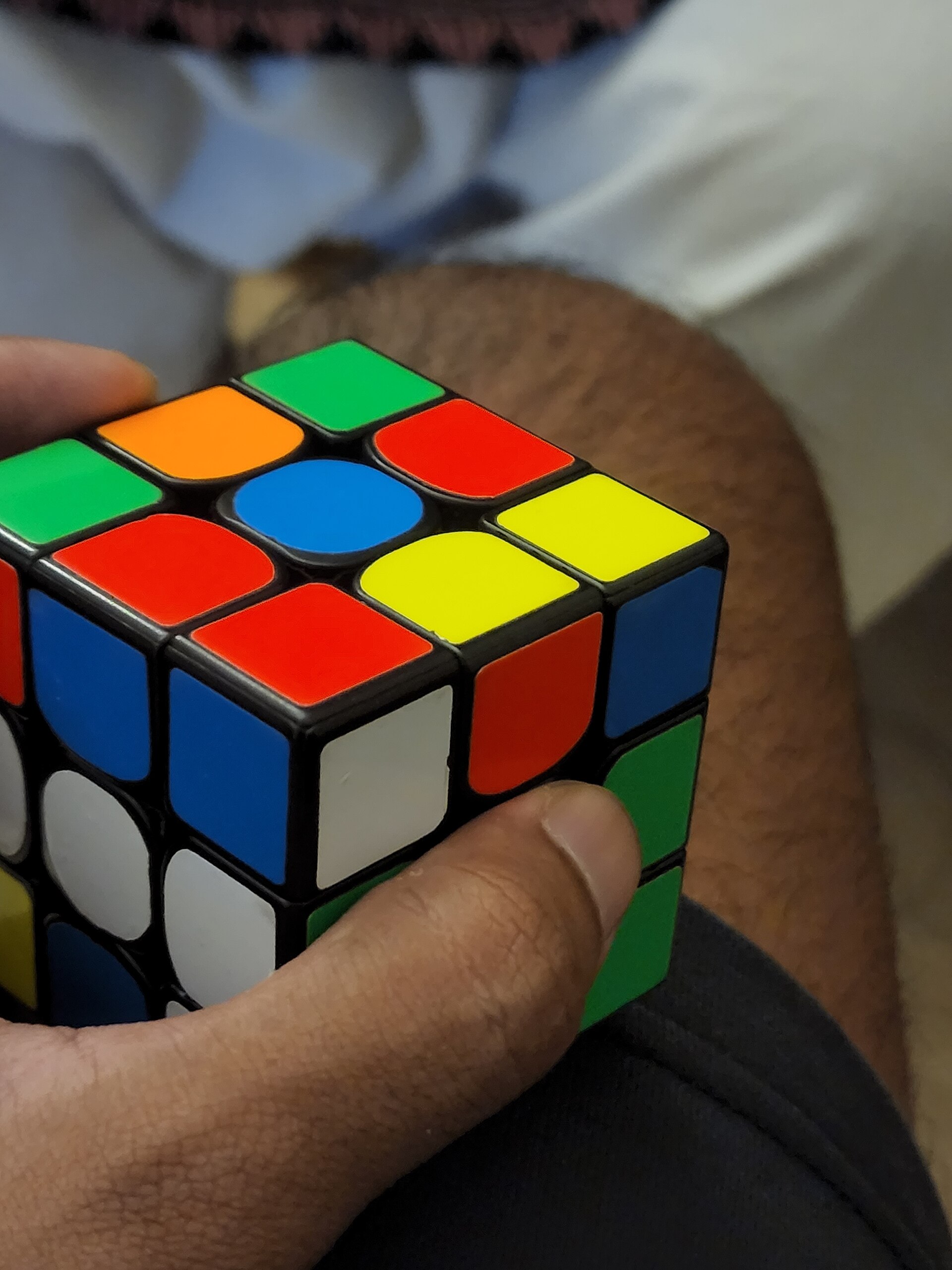 A close-up of a hand holding a scrambled three by three Rubik's Cube, the top face showing green, orange, blue, red and yellow stickers in no order